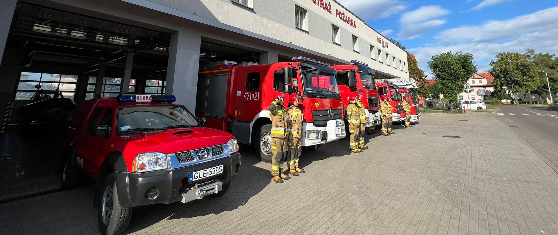 Strażacy stojący na baczność w szeregu przed budynkiem komendy. Dowódca zmiany salutuje. Samochody przed budynkiem komendy z włączonymi sygnałami świetlno-dźwiękowymi ustawione przed bramami garażowymi. 