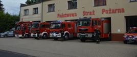79 rocznica wybuchu Powstania Warszawskiego. Na tle budynku komendy powiatowej PSP stoi 5 pojazdów pożarniczych z włączoną sygnalizacją świetlną. Obok każdego z nich stoi strażak w ubraniu koszarowym, który oddaje honor poprzez salutowanie. 