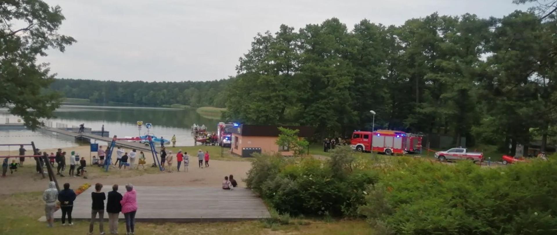 Widać ludzi na plaży, obserwują działania straży pożarnej, po prawej samochody straży
