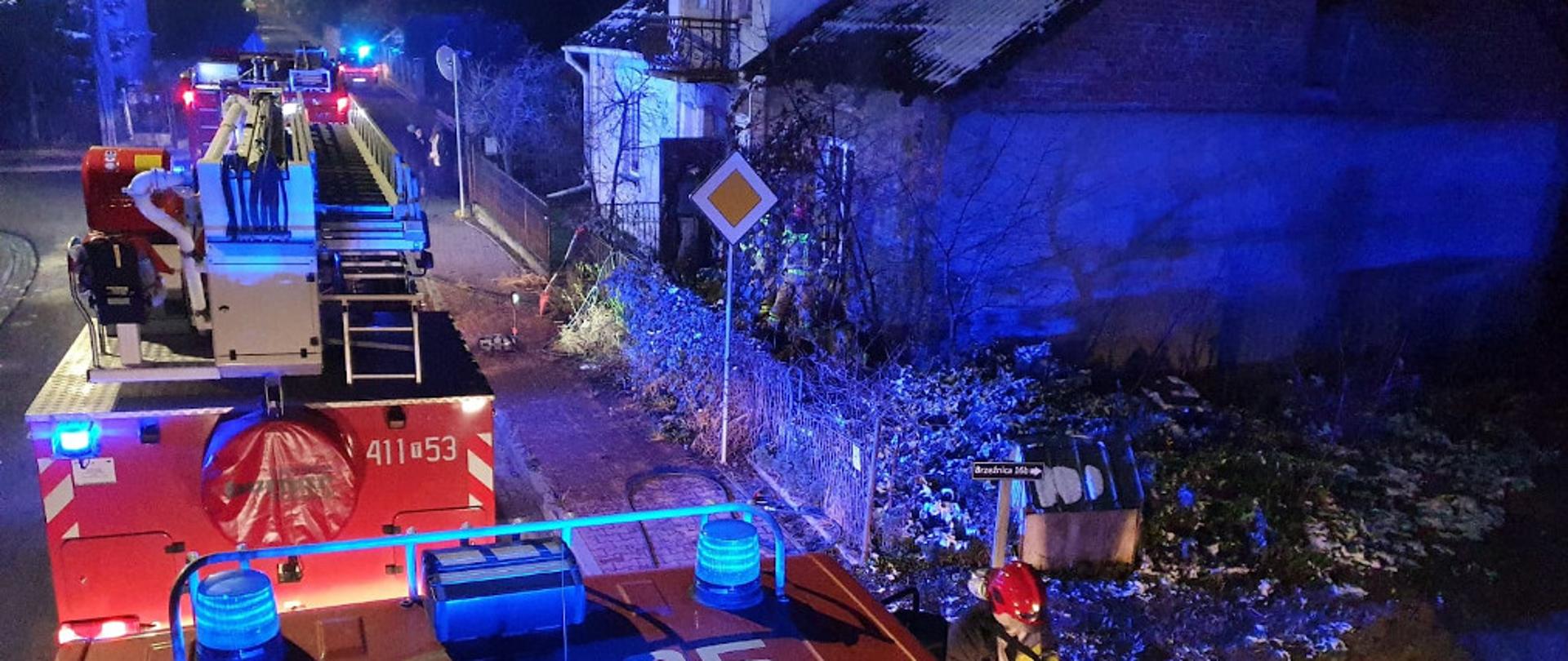 Widok na budynek z dachu samochodu gaśniczego. Na drugim planie inne pojazdy ratowniczo- gaśnicze zaparkowane na poboczu ulicy, przy której zlokalizowany jest budynek, w którym doszło do pożaru.