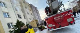 Widoczny wydobywający się dym z okien mieszkania na pierwszym piętrze. Osoby ewakuowane znajdują się przed budynkiem. Strażacy udzielają pomocy medycznej jednemu z poszkodowanych. Na ulicy sprawiany jest podnośnik hydrauliczny.