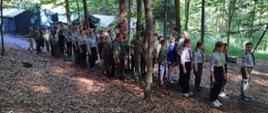 Widok z boku. Pododdział harcerski stoi w kolumnie dwójkowej wśród drzew. Za nimi w tle widoczne namioty.