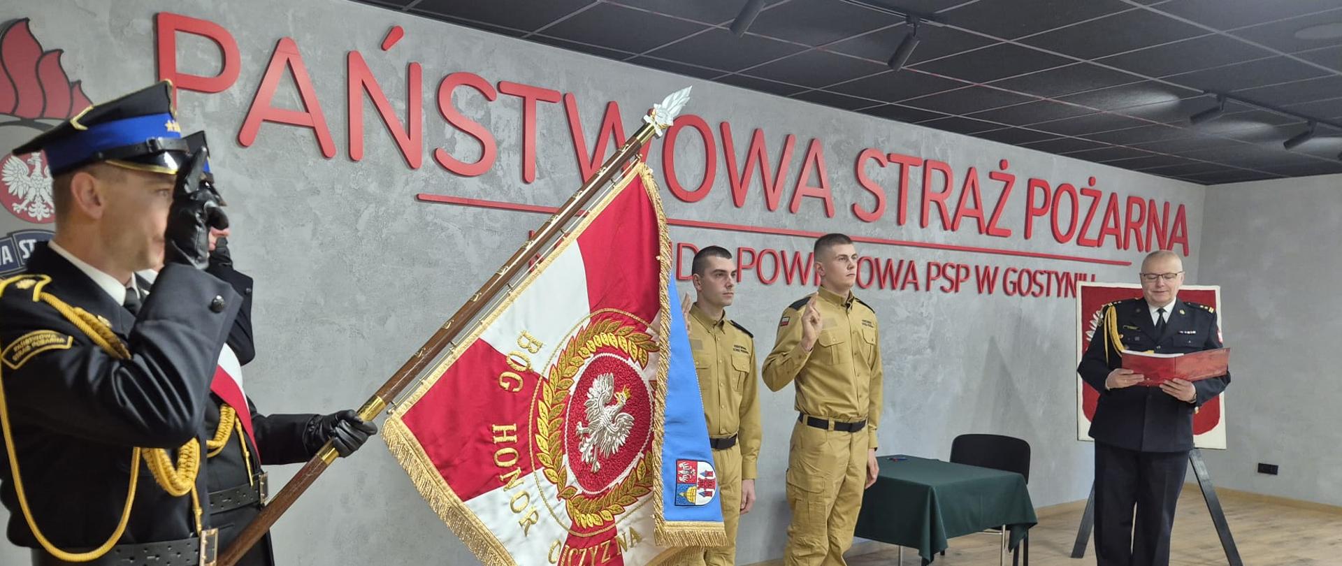 Funkcjonariuszy podczas ślubowania, którzy znajdują się w pomieszczeniu sali konferencyjnej. Dwóch, którzy ślubują stoją obok siebie na środku sali i mają podniesioną prawą rękę do góry. Z lewej strony stoi poczet sztandarowy, w którego skład wchodzi trzech funkcjonariuszy. Sztandar jest pochylony, a strażacy oddają honor poprzez salutowanie. Z prawej strony stoi komendant.