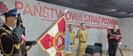 Funkcjonariuszy podczas ślubowania, którzy znajdują się w pomieszczeniu sali konferencyjnej. Dwóch, którzy ślubują stoją obok siebie na środku sali i mają podniesioną prawą rękę do góry. Z lewej strony stoi poczet sztandarowy, w którego skład wchodzi trzech funkcjonariuszy. Sztandar jest pochylony, a strażacy oddają honor poprzez salutowanie. Z prawej strony stoi komendant.