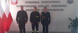 Trzech strażaków stoi w szeregu obok siebie. W tle napis na ścianie "Komenda Powiatowa Państwowej Straży Pożarnej w Makowie Mazowieckim" oraz godło Polski i logo PSP.