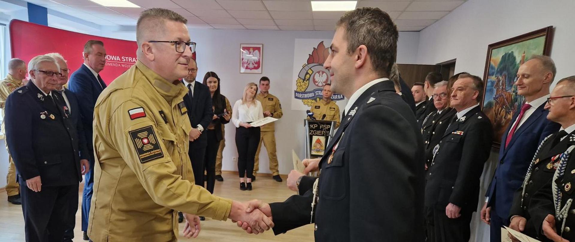 zastępca komendanta głównego psp gratuluje druhowi osp, na sali grupa druhów ochotniczych straży pożarnych w umundurowaniu galowym bez nakrycia głowy, po lewej stronie zdjęcia przedstawiciele rządu oraz samorząd terytorialny, na ścianie wisi godło państwowe oraz logo psp
