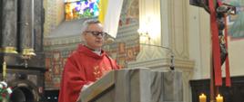 Zdjęcie przedstawia postać księdza ubranego w czerwone szaty przemawiającego z ambony podczas mszy świętej. Po prawej stronie widoczny krzyż.