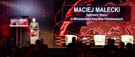 Wiceminister Maciej Małecki przemawia z mównicy. Przed nim siedzą słuchacze na krzesełkach. W tle ekran z logo konferencji i imieniem i nazwiskiem ministra.