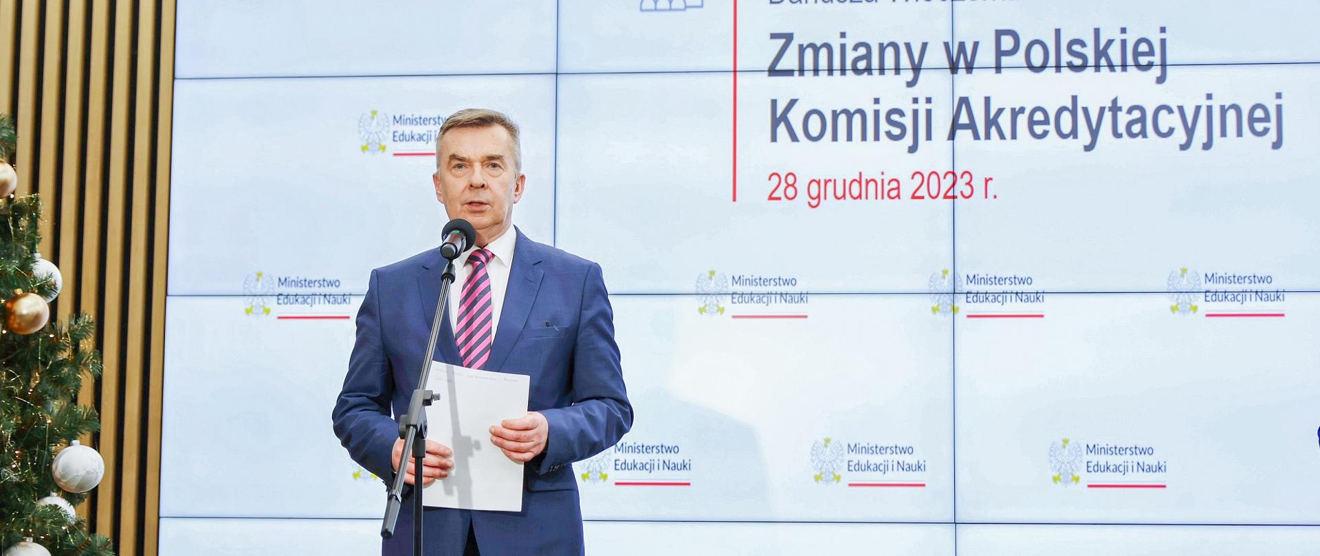 Minister Wieczorek stoi i mówi do mikrofonu na stojaku, za nim wbudowany w ścianę wieli ekran z napisem Zmiany w Polskiej Komisji Akredytacyjnej 28 grudnia 2023 r.