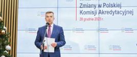 Minister Wieczorek stoi i mówi do mikrofonu na stojaku, za nim wbudowany w ścianę wieli ekran z napisem Zmiany w Polskiej Komisji Akredytacyjnej 28 grudnia 2023 r.