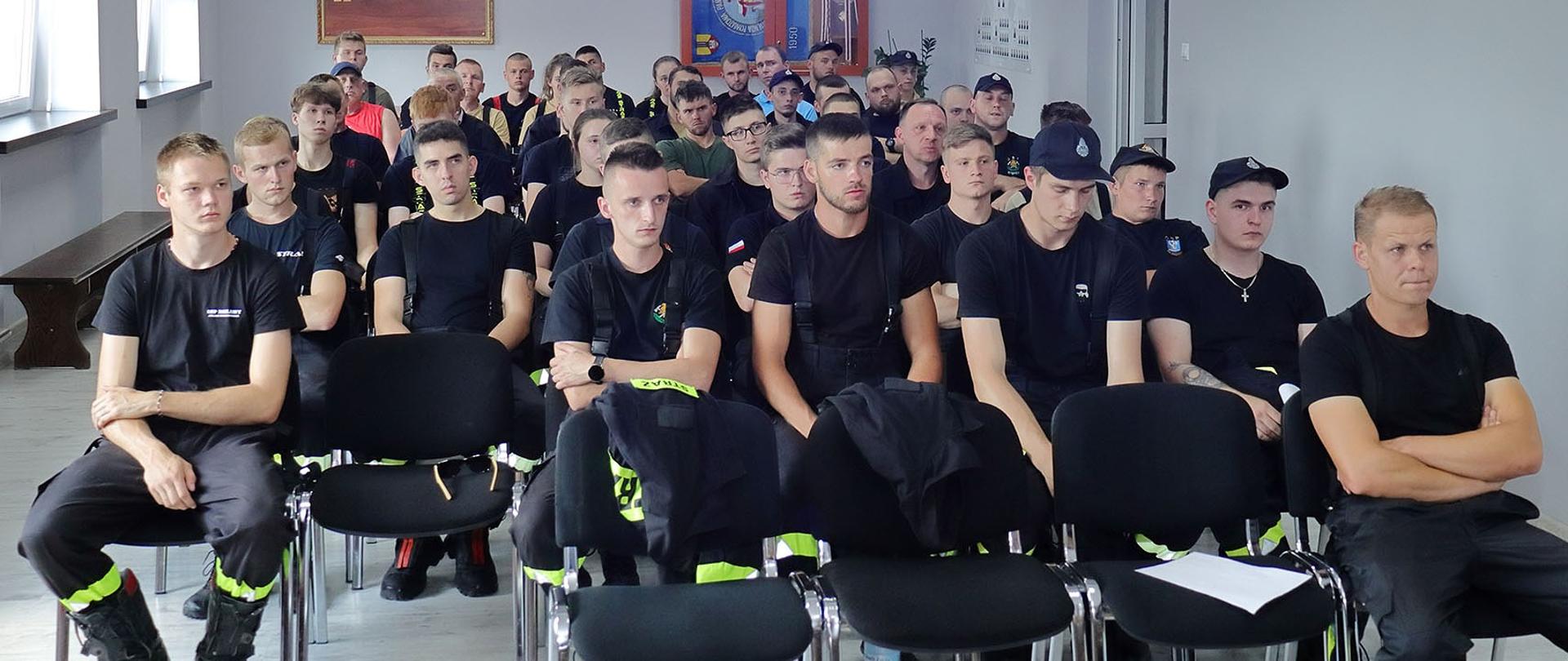 kilkadziesiąt osób w umundurowaniu koszarowym osp siedzi na krzesłach w tle sztandar w gablocie i drewniana tablica pamiątkowa 