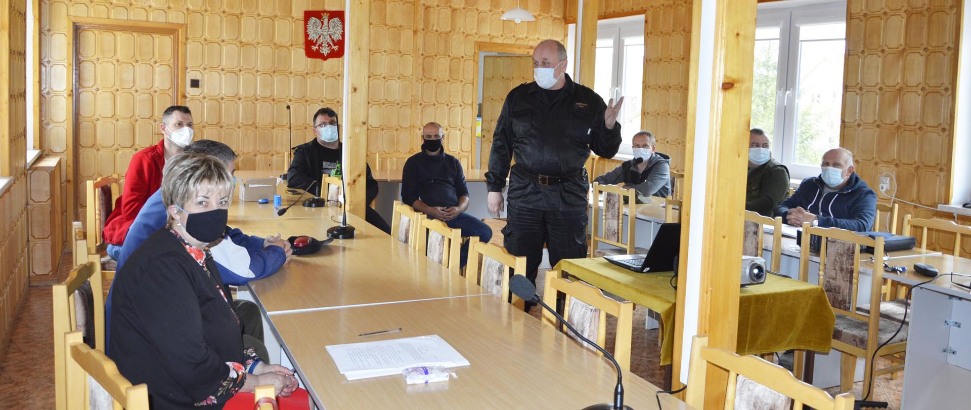 Na zdjęciu widać zastępce komendanta powiatowego PSP w Ustrzykach Dolnych w ubraniu dowódczo-sztabowym który stoi na środku pomieszczenia przy stoliku na którym stoi laptop i rzutnik. W około stoją stoły a przy nich siedzi osiem osób w ubraniach cywilnych. Wszyscy mają założone maseczki na twarzy.