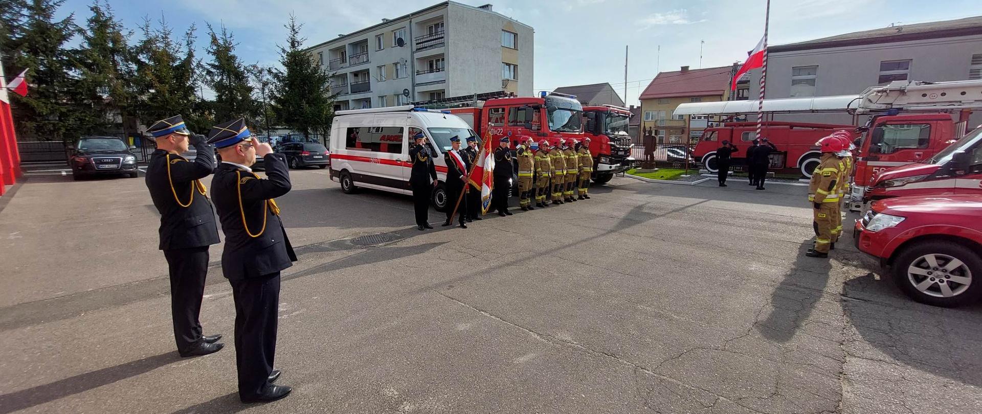 Zmiany służbowe i poczet sztandarowy stoją naprzeciwko siebie przed samochodami strażackimi na placu komendy. Pośrodku stoi Komendant Powiatowy PSP oraz dowódca JRG PSP. Poczet flagowy wciąga flagę na maszt. Dowódcy pododdziałów salutują.