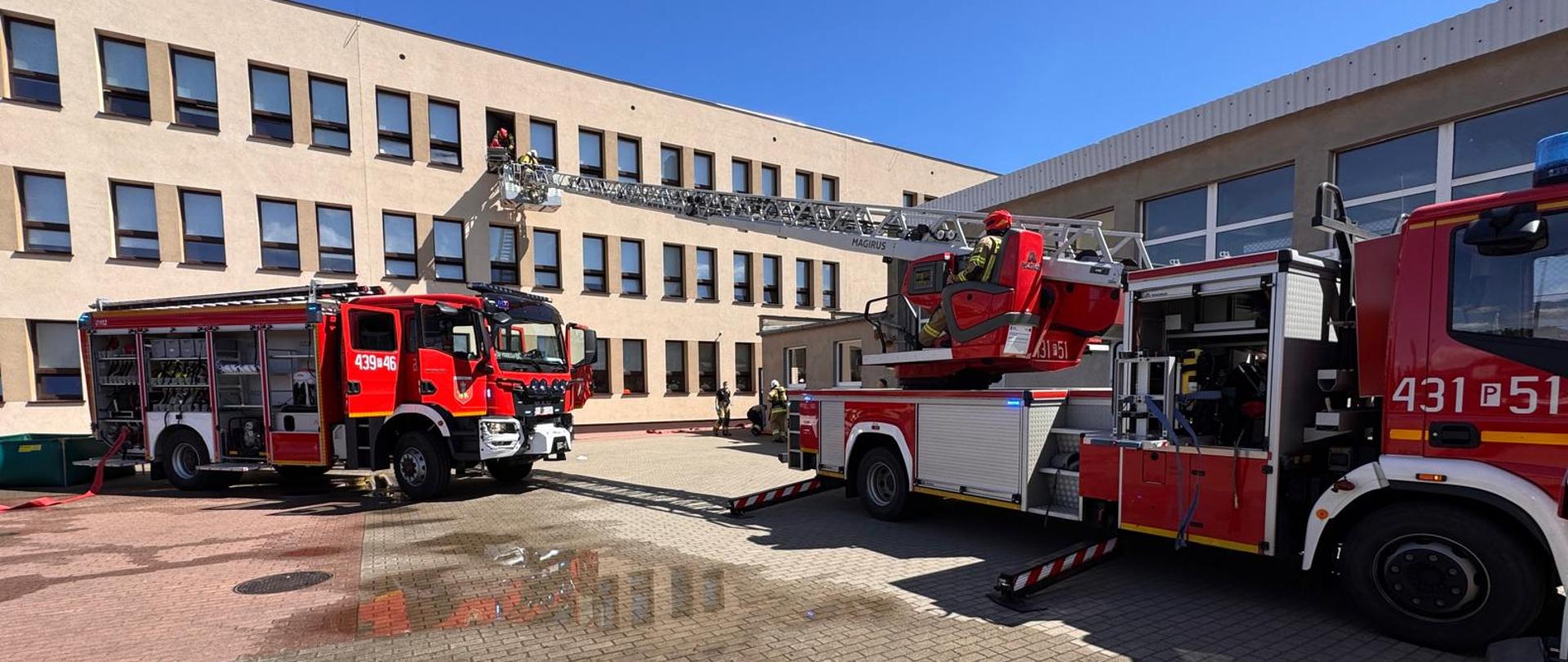 Dwa samochody strażackie stojący przy budynku szkoły. Jeden to drabina mechaniczna. Kosz drabiny podstawiony jest pod okno. Strażacy ewakuują osobę poszkodowaną za pomocą tej drabiny.