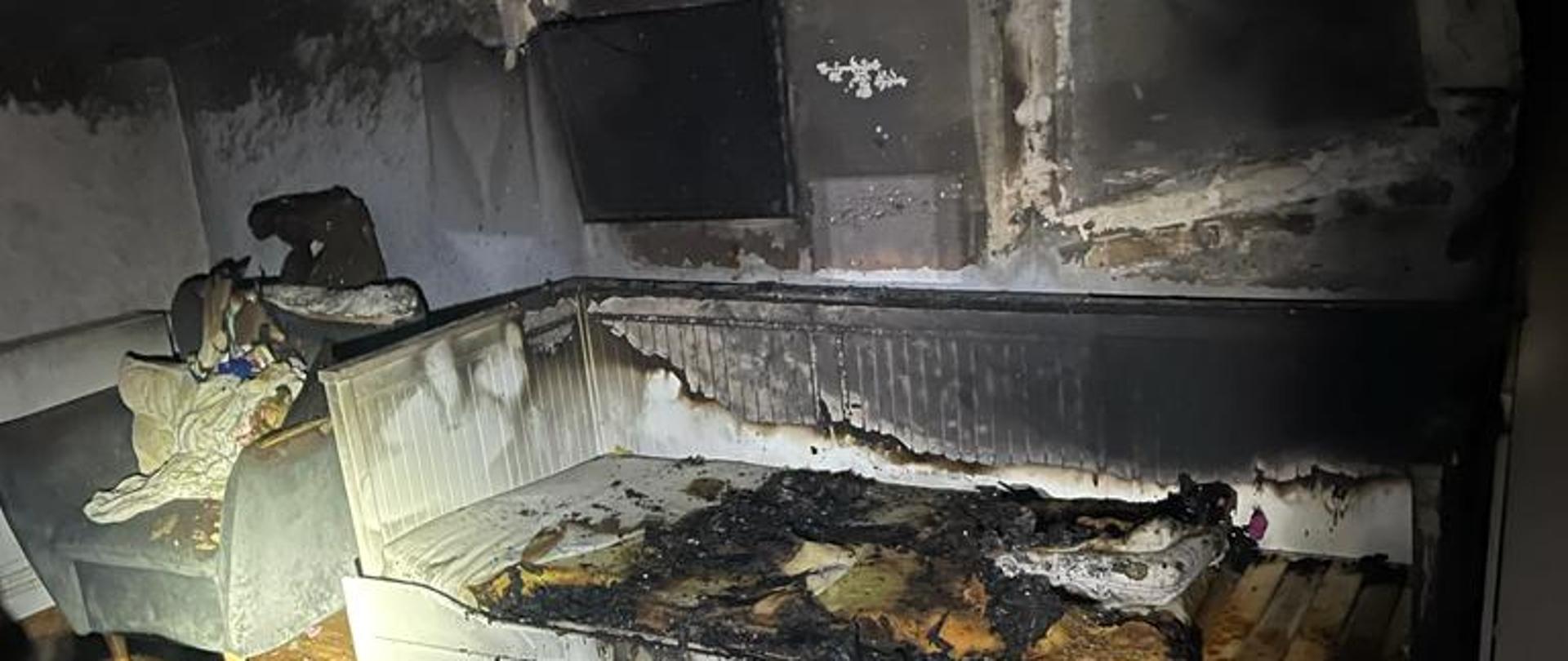 widok pokoju w mieszkaniu w którym powstał pożar. Spalone łóżko, okopcone ściany, sufit i całe wyposażenie pokoju.
