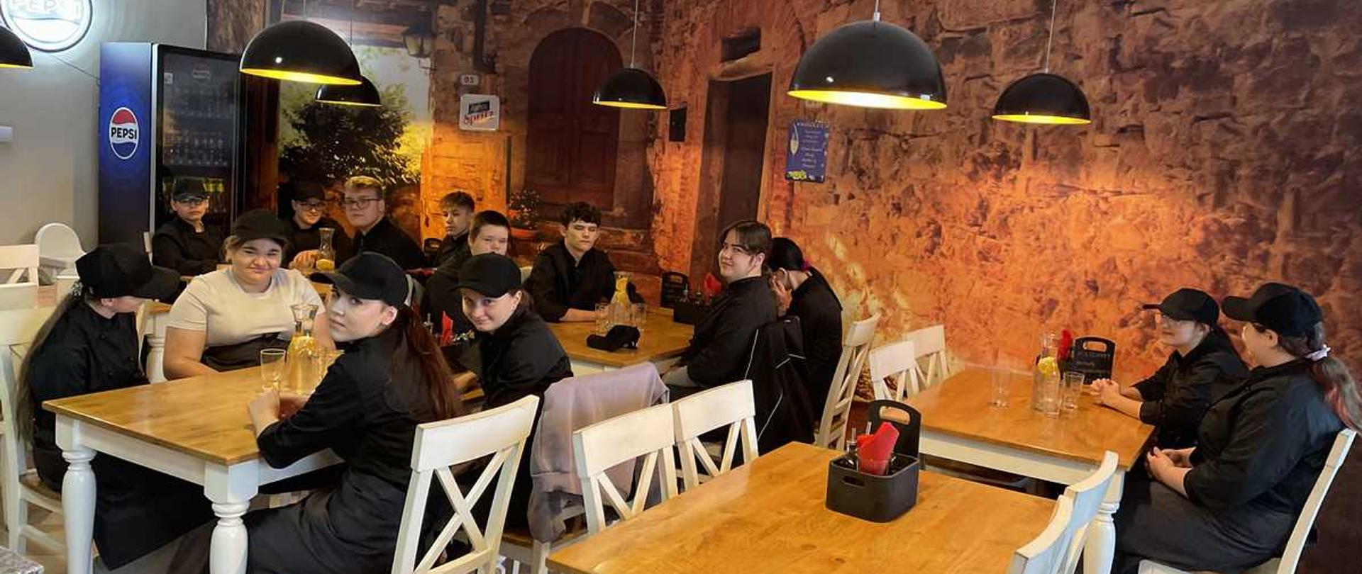 Zdjęcie przedstawia grupę młodych ludzi siedzących przy drewnianych stołach w restauracji lub kawiarni. Wszyscy mają na sobie czarne ubrania (bluzy lub kurtki) i czarne czapki z daszkiem.
Wnętrze jest jasne, z podłogą wyłożoną jasnymi płytkami. Ściana w tle jest ozdobiona dużą fototapetą imitującą stary, kamienny mur z niszami i łukami. Sufit jest biały, z kilkoma wiszącymi czarnymi lampami w kształcie kopuł oraz białymi wiatrakami sufitowymi. Po lewej stronie w tle widoczna jest lodówka z logo Pepsi.
Na stołach, które są z jasnego drewna, znajdują się białe krzesła z wyciętymi w oparciach wzorami w kształcie litery ""X"". Na stołach stoją szklane karafki z napojami i pojemniki na przyprawy, takie jak ketchup. Grupa jest podzielona na mniejsze podgrupy siedzące przy osobnych stołach, a ich uwaga wydaje się być skierowana w różne strony, niektórzy patrzą w stronę aparatu, inni rozmawiają między sobą.