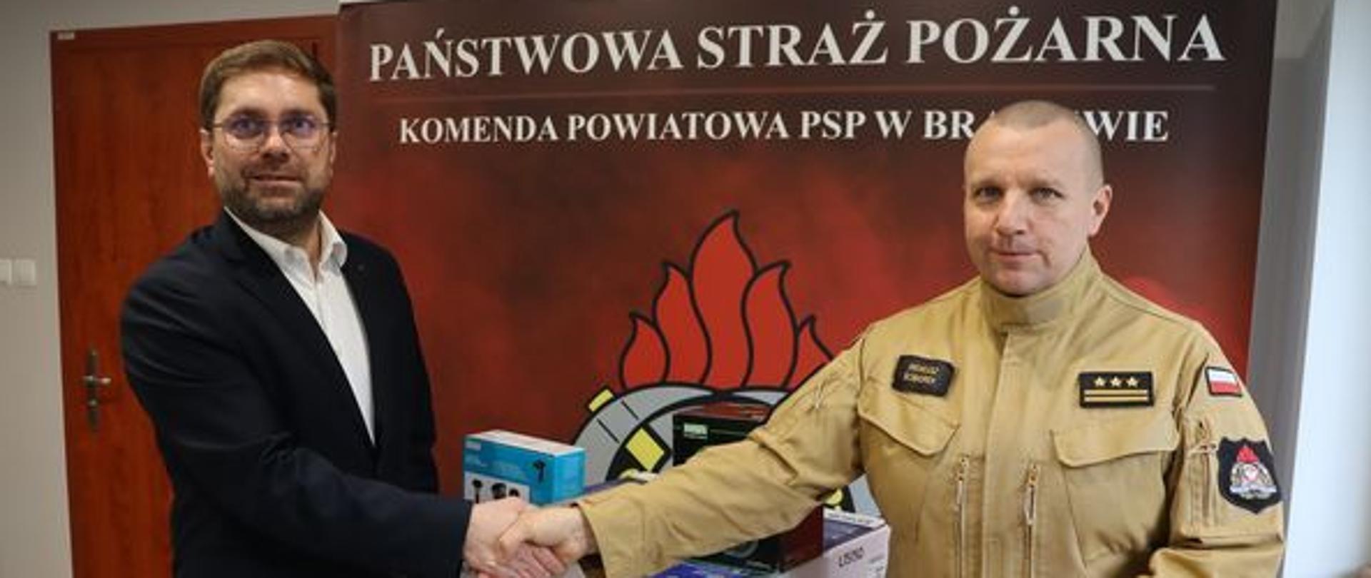 Wójt gminy Braniewo z lewej ściska dłoń komendantowi powiatowemu. Na dole przekazany sprzęt, duże pudła. W tle baner Państwowa straż pożarna.