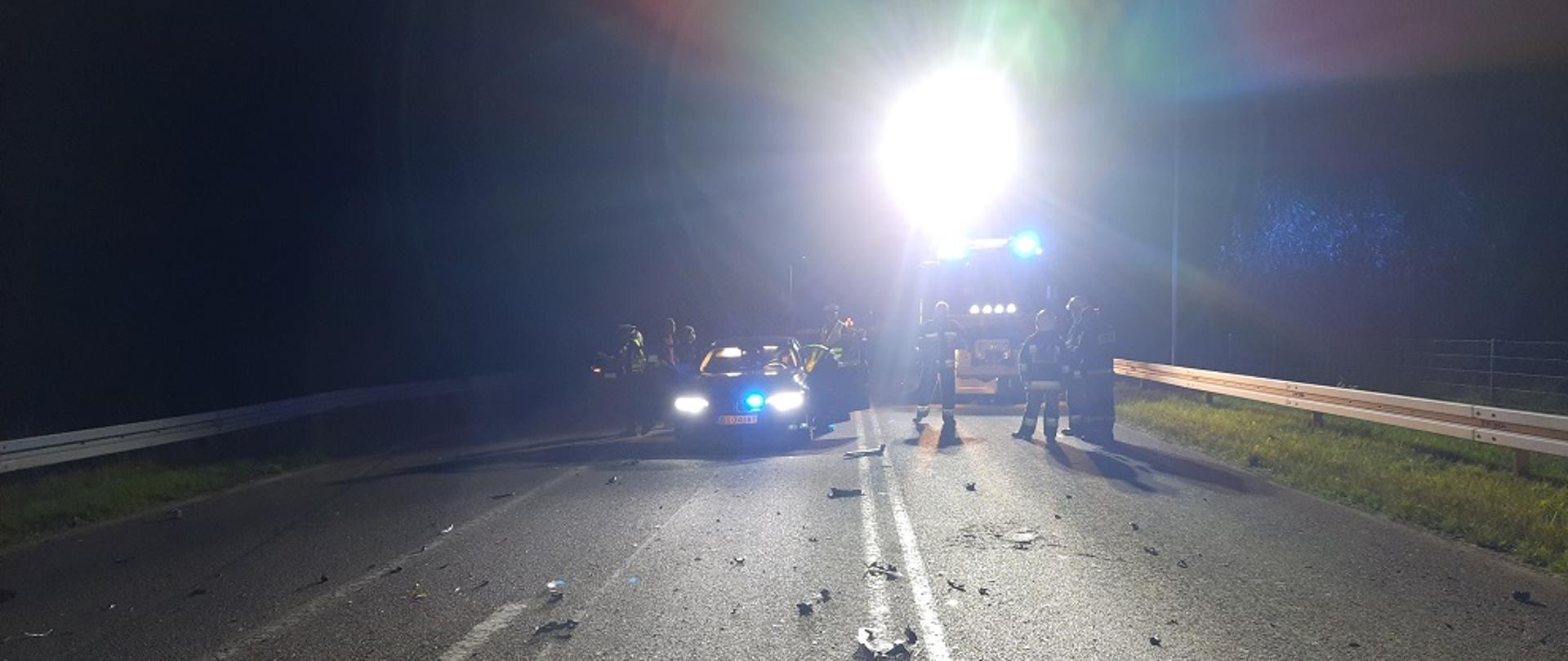 Samochody straży stoją na włączonych światłach. Jasne, oslepiające światło z masztu oświetleniowego skierowane w stronę obiektywu. Na jezdni porozrzucane części plastikowe z pojazdu. Dookoła panuje zmrok.