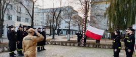 Strażacy stoją w rzędach naprzeciwko siebie poczet sztandarowy wiesza flagę na masz strażak w mundurze musztardowym stoi przed rzędami oraz flagą oddając honor.