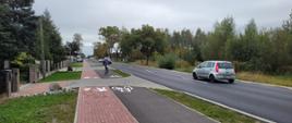 Ciąg pieszo‑rowerowy z wydzielonymi pasami dla pieszych i rowerzystów biegnący wzdłuż drogi, po której poruszają się samochody. W pobliżu znajdują się ogrodzenia posesji, drzewa i niska zabudowa.
