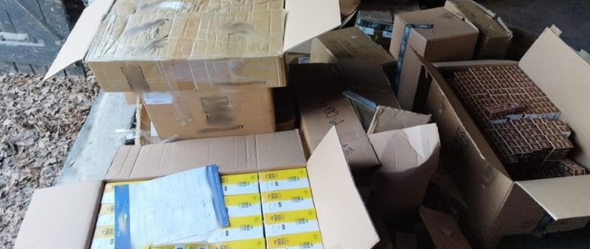 Na podłodze ustawione są liczne kartony zawierające wyroby tytoniowe i materiały opakowaniowe, częściowo otwarte i ułożone nieregularnie.