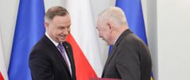Uśmiechnięty prezydent Duda ściska rękę siwemu mężczyźnie w garniturze, który trzyma czerwoną teczkę. Za nimi flagi Polski i UE.