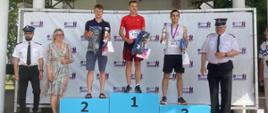 stojący na podium trzej zawodnicy w strojach sportowych z medalami, po bokach podium stoją ubrani w białe koszule i czapki rogatywki strażacy