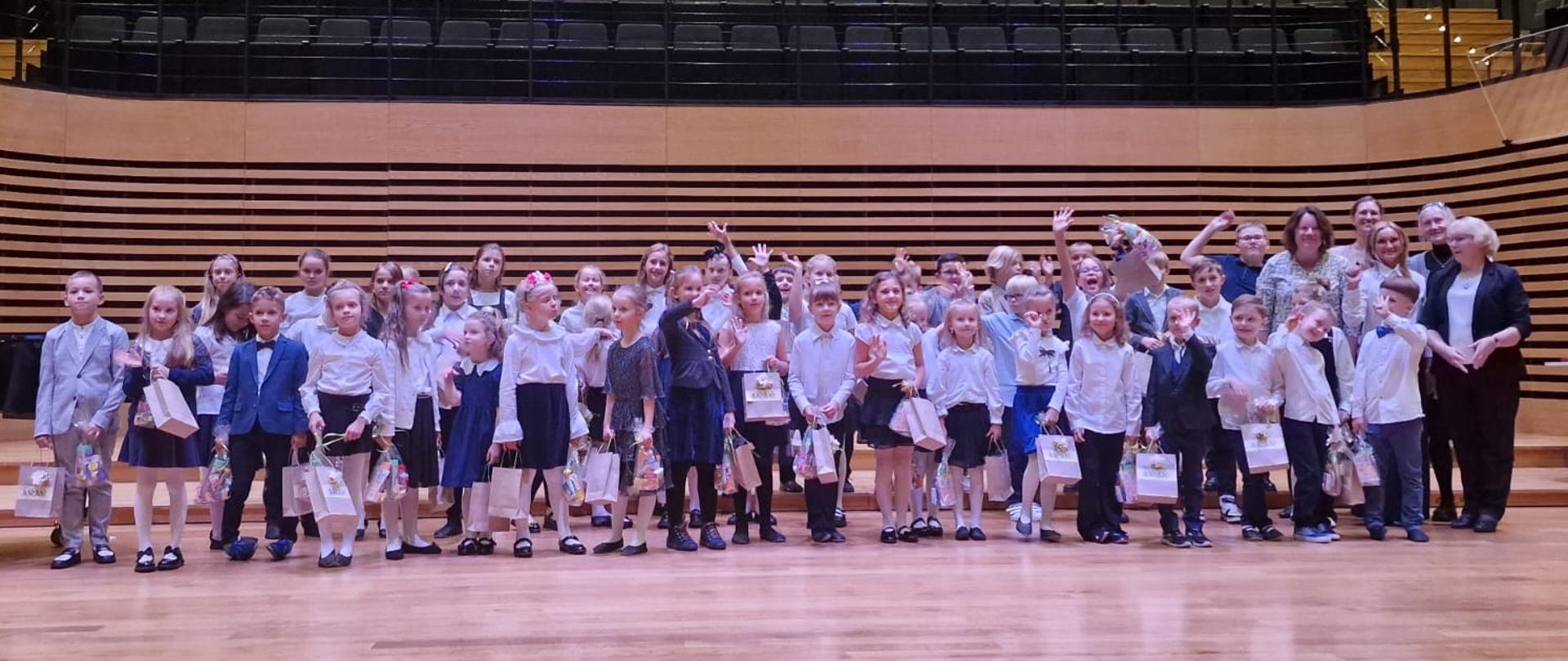 Wszyscy uczniowie klas pierwszych stoją na scenie w dwóch rzędach, za nimi z boku stoi pięć nauczycielek.