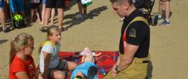 Piknik "Kręci mnie bezpieczeństwo nad wodą". Na kocu 3 manekiny do nauki zasad udzielania pierwszej pomocy. Z lewej strony klęczą dwie dziewczynki, a na przeciwko nich strażak instruujący w jaki sposób prowadzić zewnętrzny masaż serca i sztuczne oddychanie. W tle pozostali uczestnicy pikniku.