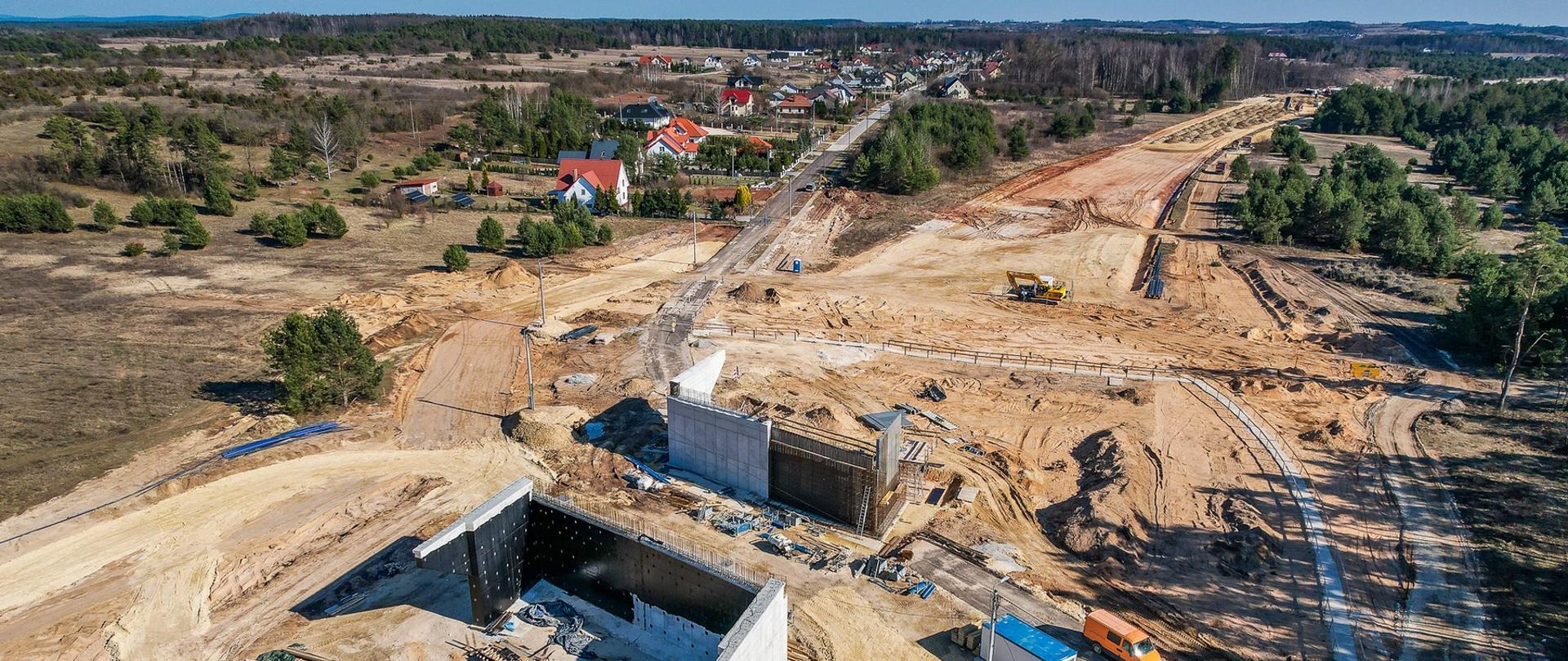 Budowa obwodnicy Morawicy - zdjęcie budowy wykonane z góry, na pierwszym planie dwie betonowe podpory wiaduktu, dalej koparka, piaszczysty ślad przyszłej drogi, w oddali domy jednorodzinne 