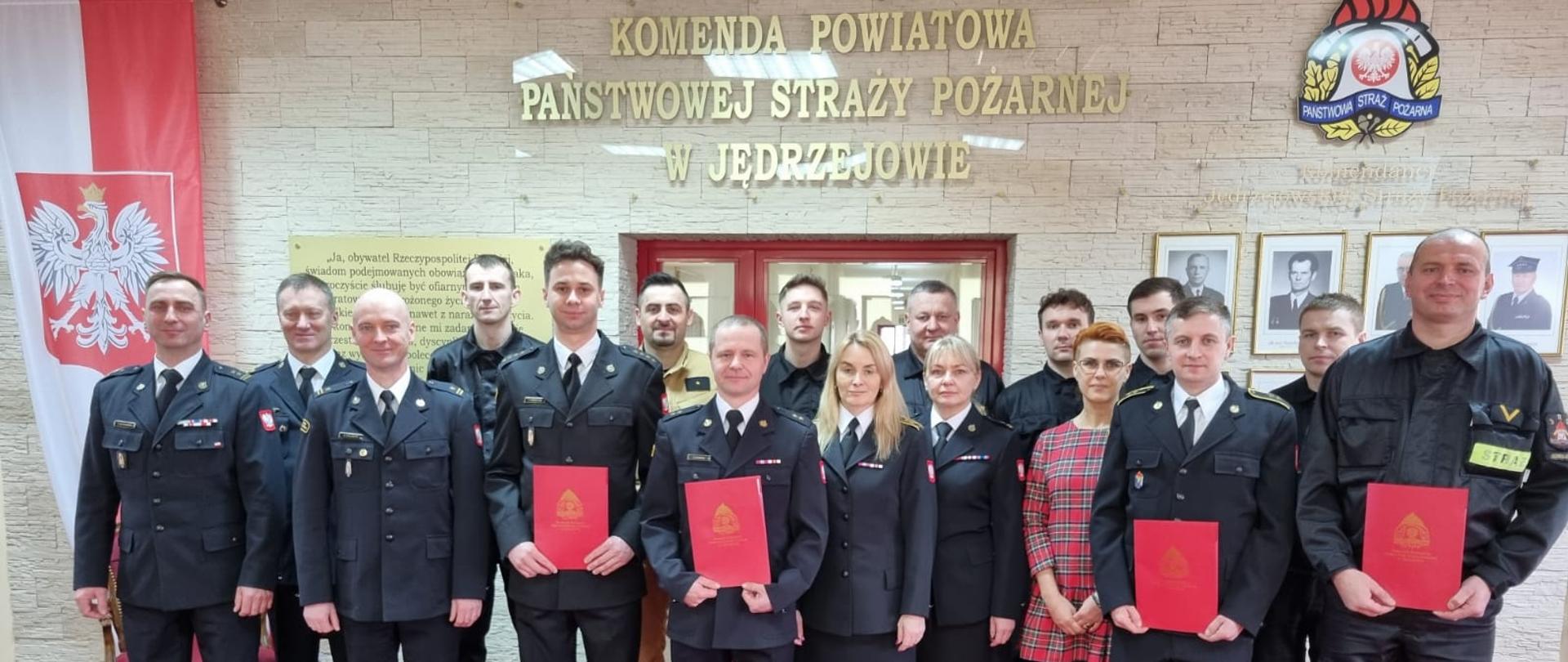 Zdjęcie przedstawia strażaków oraz pracowników cywilnych Komendy Powiatowej Państwowej Straży Pożarnej w Jędrzejowie podczas uroczystej zbiórki z okazji nadania wyższych stopni służbowych. Zdjęcie zrobione w sali tradycji tutejszej komendy.