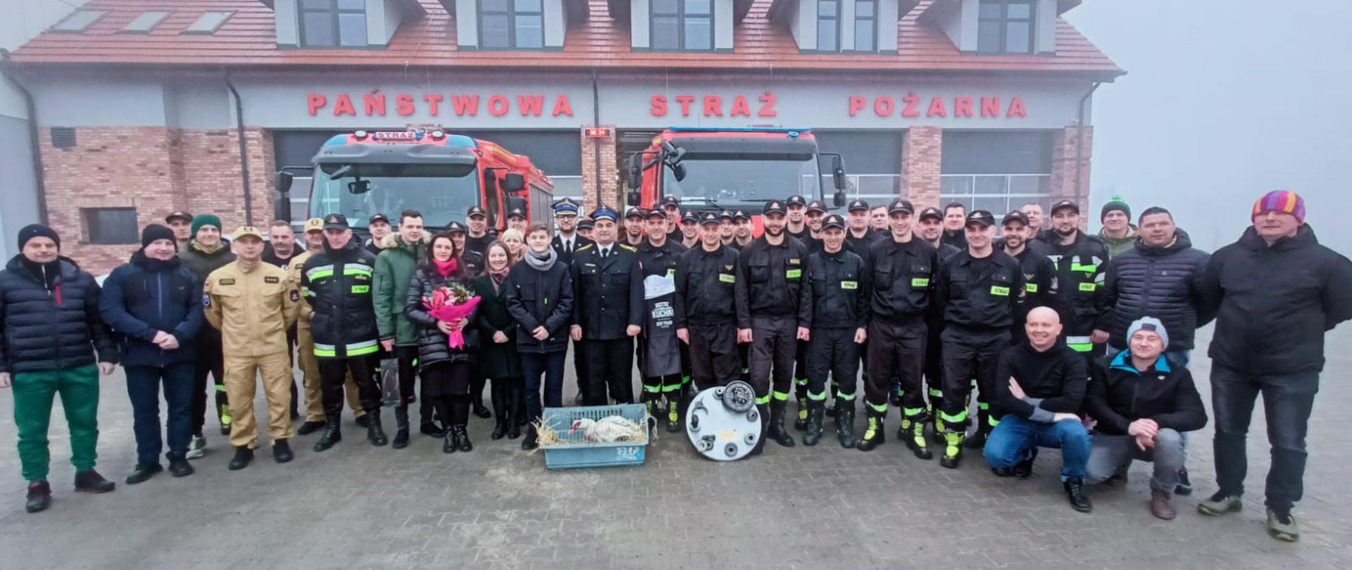 Przed budynkiem Komendy Powiatowej Państwowej Straży Pożarnej w Świeciu na tle samochodów pożarniczych stoją uczestnicy pożegnania ze służba st. ogn. Patryka Kaczerowskiego.