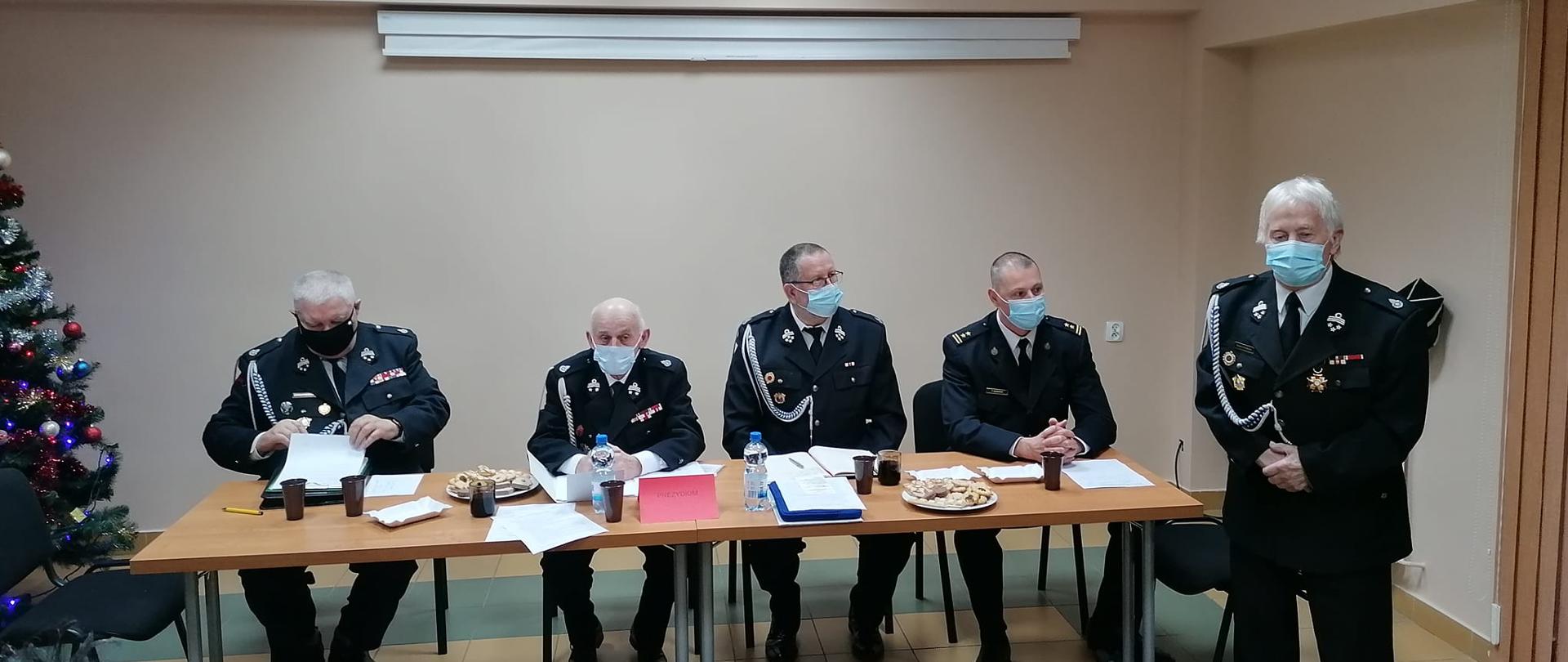 Oświetlone pomieszczenie, przy stole siedzą od prawej trzej druhowie w mundurach oraz komendant PSP w Sztumie, po prawej stronie stoi umundurowany druh, wszyscy są w maseczkach ochronnych na twarzach. Po lewej stronie kadru widać kawałek ustrojonej choinki.