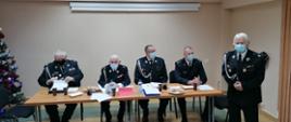 Oświetlone pomieszczenie, przy stole siedzą od prawej trzej druhowie w mundurach oraz komendant PSP w Sztumie, po prawej stronie stoi umundurowany druh, wszyscy są w maseczkach ochronnych na twarzach. Po lewej stronie kadru widać kawałek ustrojonej choinki.