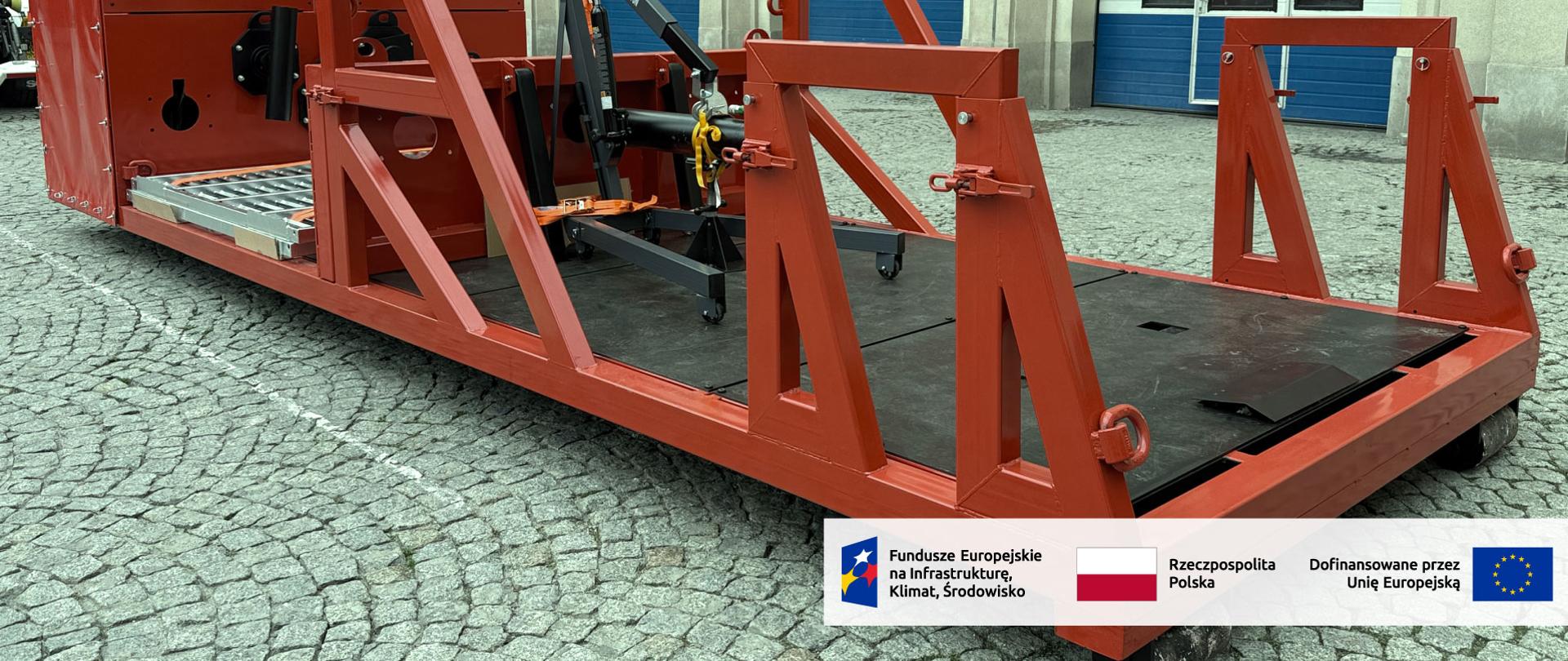 Czerwona stalowa platforma z ramą konstrukcyjną i zamontowanym podnośnikiem, ustawiona na utwardzonym placu przed garażami; na konstrukcji widoczne logo producenta oraz oznaczenia projektu współfinansowanego przez Unię Europejską.
