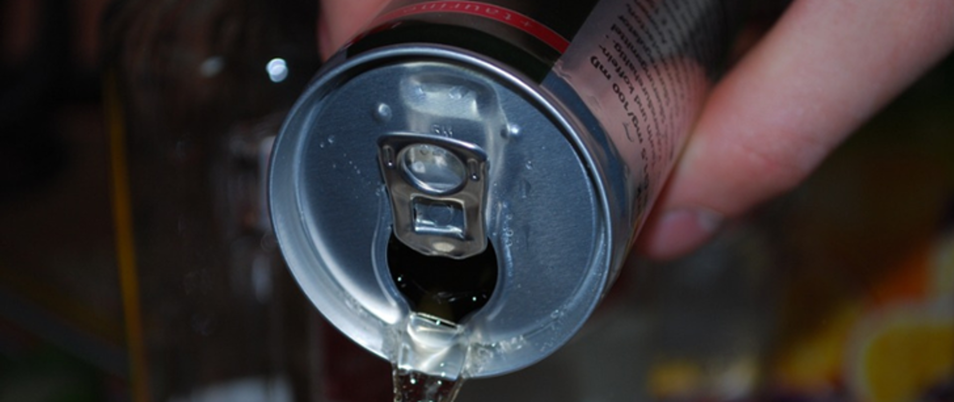 Na zdjęciu znajduje się puszka trzymana w dłoni, z której wylewa się ciecz.