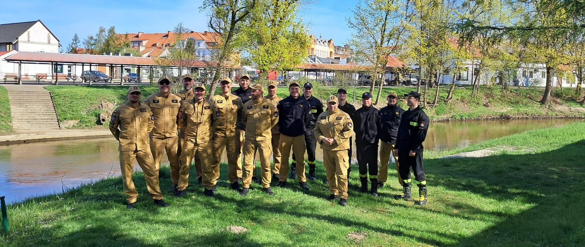 Nad brzegiem rzeki stoi grupa strażaków. Słonecznie.