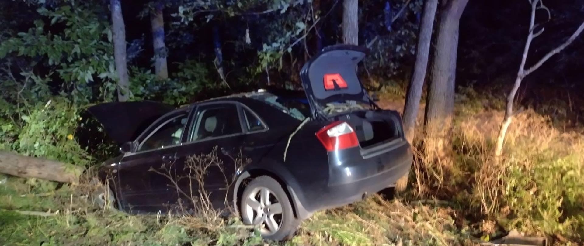 Na zdjęciu widać uszkodzony pojazd osobowy na poboczu drogi. Pora nocna. Oświetlenie sztuczne. 