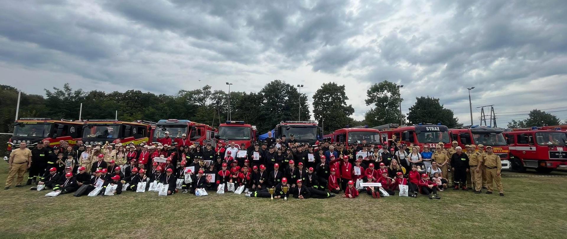 Zdjęcie przedstawia drużyny ustawione na tle samochodów strażackich.
