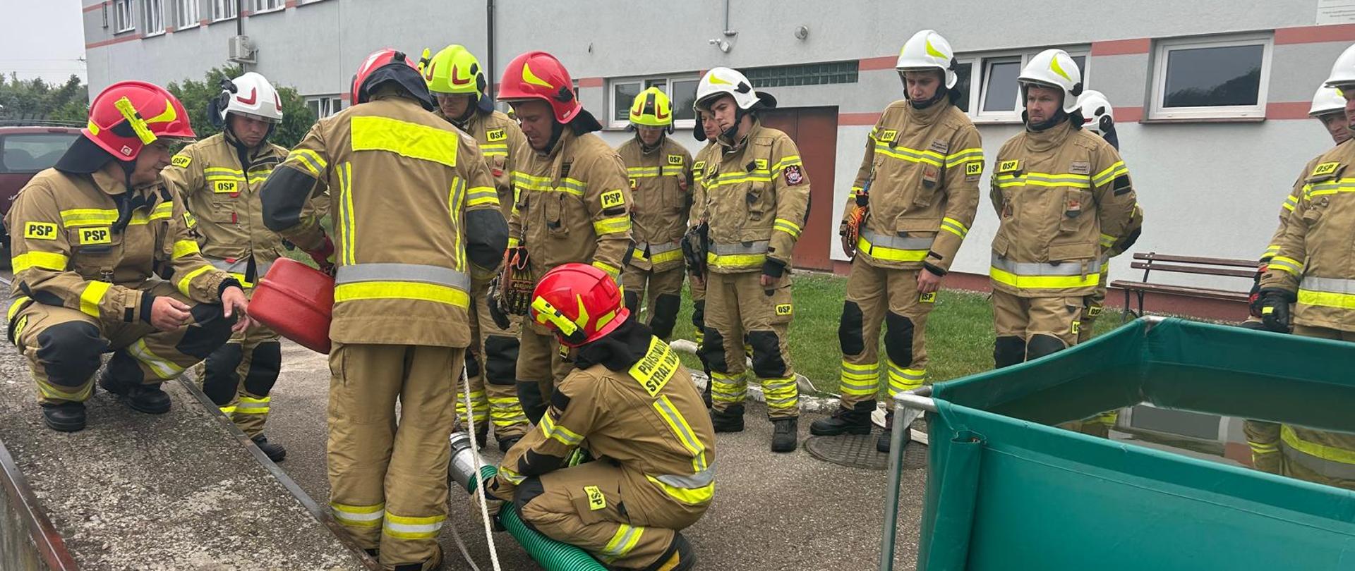 Grupa strażaków w umundurowaniu koloru piaskowego stoi na placu na tle szarego budynku z prawej strony swoi zielony zbiornik z wodą i zielony wąż który jeden ze strażaków trzyma