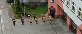 Zdjęcie przedstawia 10 strażaków w ubraniach specjalnych stojących na baczność. Widok z góry.