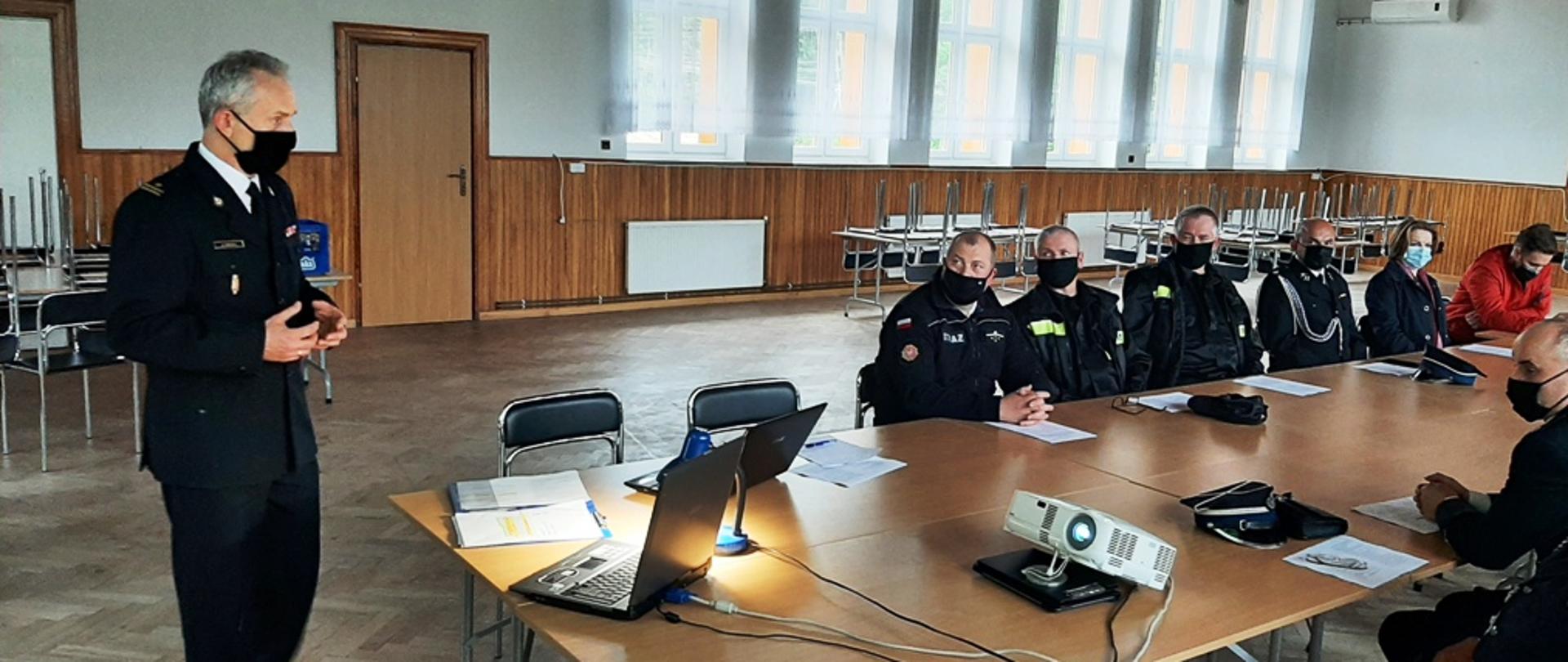 Kolorowa fotografia przedstawia wnętrze dużej sali w budynku. Na pierwszym planie widoczna postać stojącego funkcjonariusza Państwowej Straży Pożarnej w białej koszuli i czarnym mundurze galowym z dystynkcjami, w maseczce na twarzy, zwróconego do siedzących na sali. Po prawej stronie widoczne siedzące przy stolikach osoby w mundurach strażackich ochotniczej straży pożarnej, w maseczkach na twarzach. Również widoczne osoby cywilne siedzące obok. W tle okna sali remizy strażackiej. Na stoliku widoczny sprzęt do prezentacji (laptop, rzutnik).