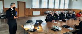 Kolorowa fotografia przedstawia wnętrze dużej sali w budynku. Na pierwszym planie widoczna postać stojącego funkcjonariusza Państwowej Straży Pożarnej w białej koszuli i czarnym mundurze galowym z dystynkcjami, w maseczce na twarzy, zwróconego do siedzących na sali. Po prawej stronie widoczne siedzące przy stolikach osoby w mundurach strażackich ochotniczej straży pożarnej, w maseczkach na twarzach. Również widoczne osoby cywilne siedzące obok. W tle okna sali remizy strażackiej. Na stoliku widoczny sprzęt do prezentacji (laptop, rzutnik).