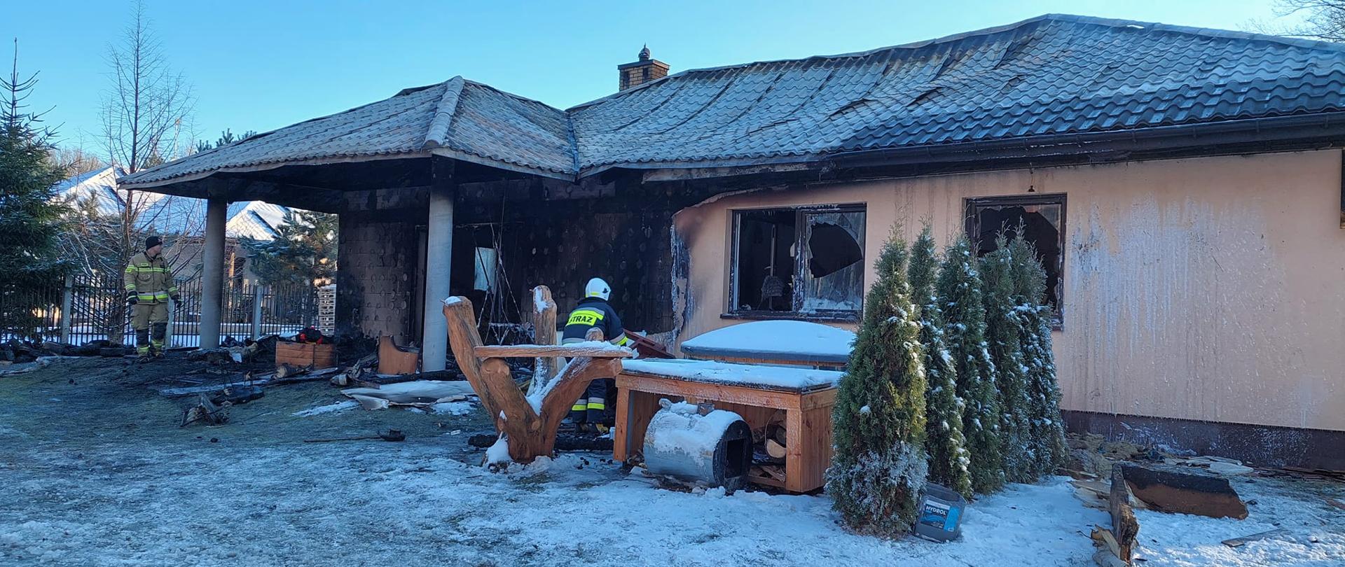 Pracujący strażacy na zewnatrz zniszczonego w pożarze domu jednorodzinnego. Widać zniszczona i pogiętą blachę pokrycia dachu, zniszczone w skutek temperatury okna i częściowo spaloną elewację budynku.