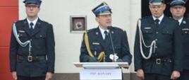 Jubileusz 95-lecia OSP Konary. Plac przed remizą. Na tle pamiątkowej tablicy, przy mównicy stoi komendant powiatowy PSP w Rawiczu, który składa gratulacje i życzenia druhom z Konar. 