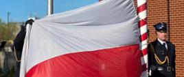 Dwóch strażaków w galowych mundurach rozwija dużą biało-czerwoną flagę przy czerwono-białym maszcie, przygotowując ją do podniesienia. Obok stoi trzeci strażak na baczność. W tle widoczny jest ceglany budynek, trawnik oraz drugi, pusty maszt flagowy. Na dole znajduje się emblemat Państwowej Straży Pożarnej z napisem „Gryfice”.