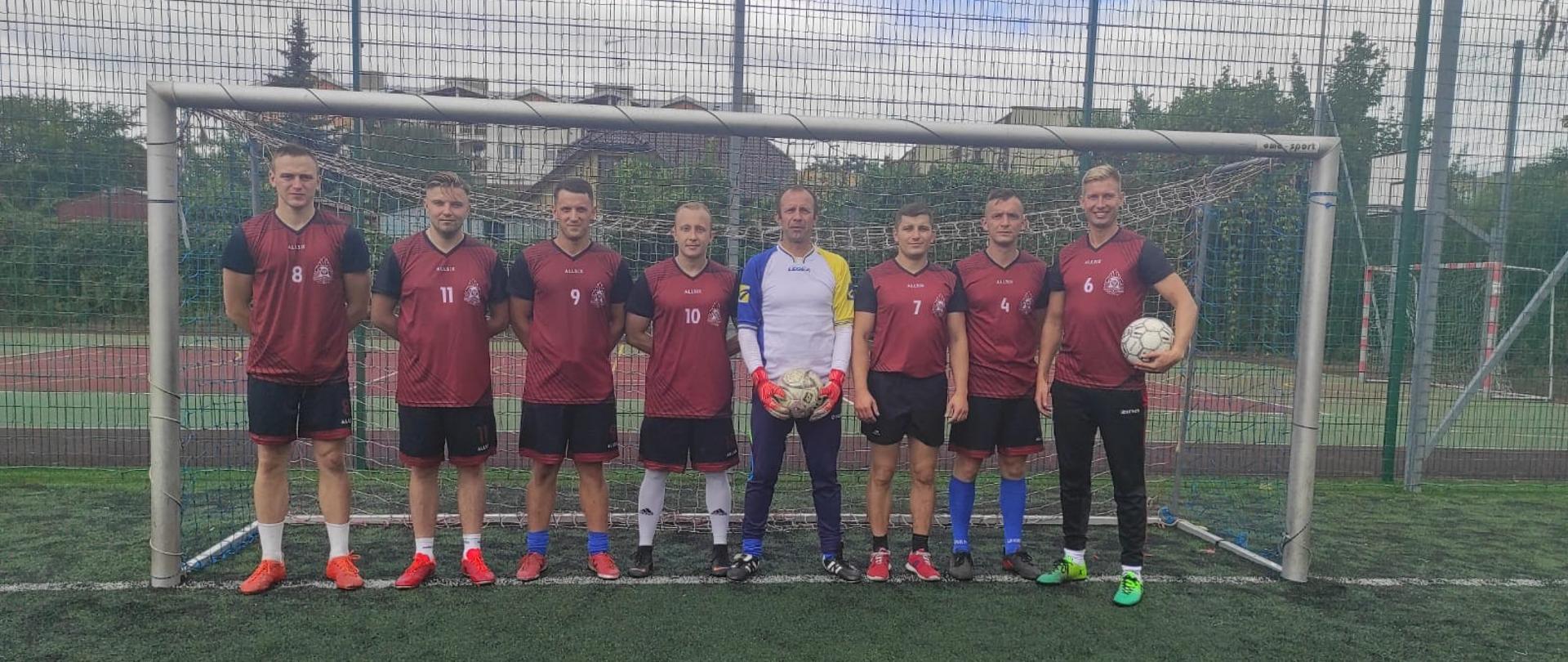 Na zdjęciu w bramce na boisku do piłki nożnej ustawiona jest drużyna z KP PSP Wołomin w czerwono - czarnych strojach. W tle widać drzewa i zabudowania. 