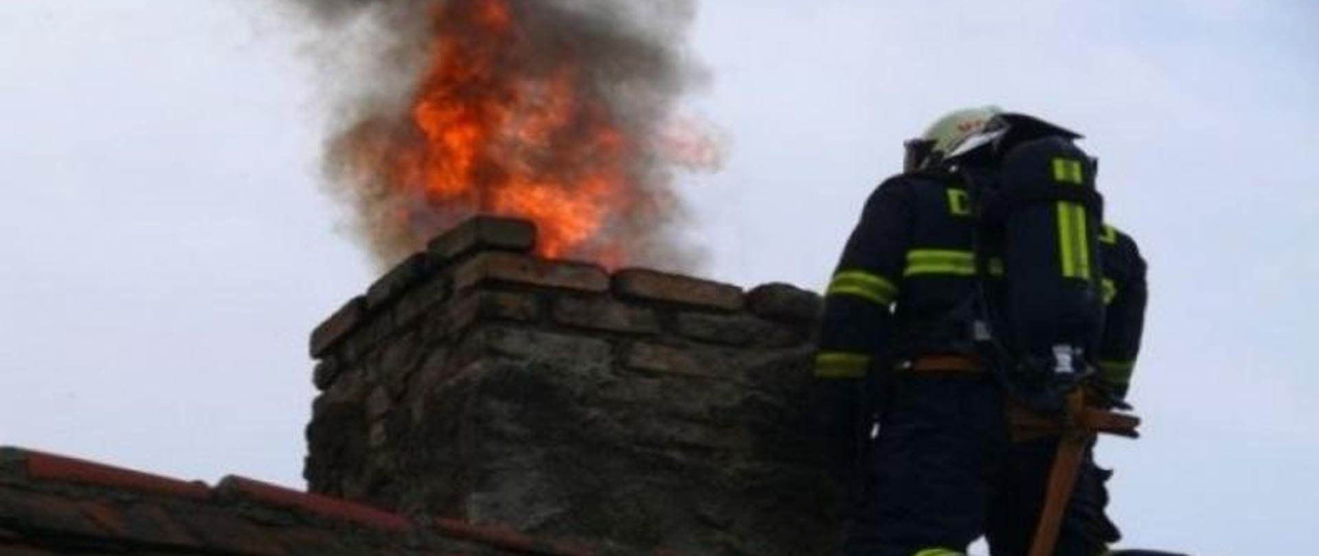 Na zdjęciu widać strażaka przy kominie z którego wydobywają się płomienie i czarny dym