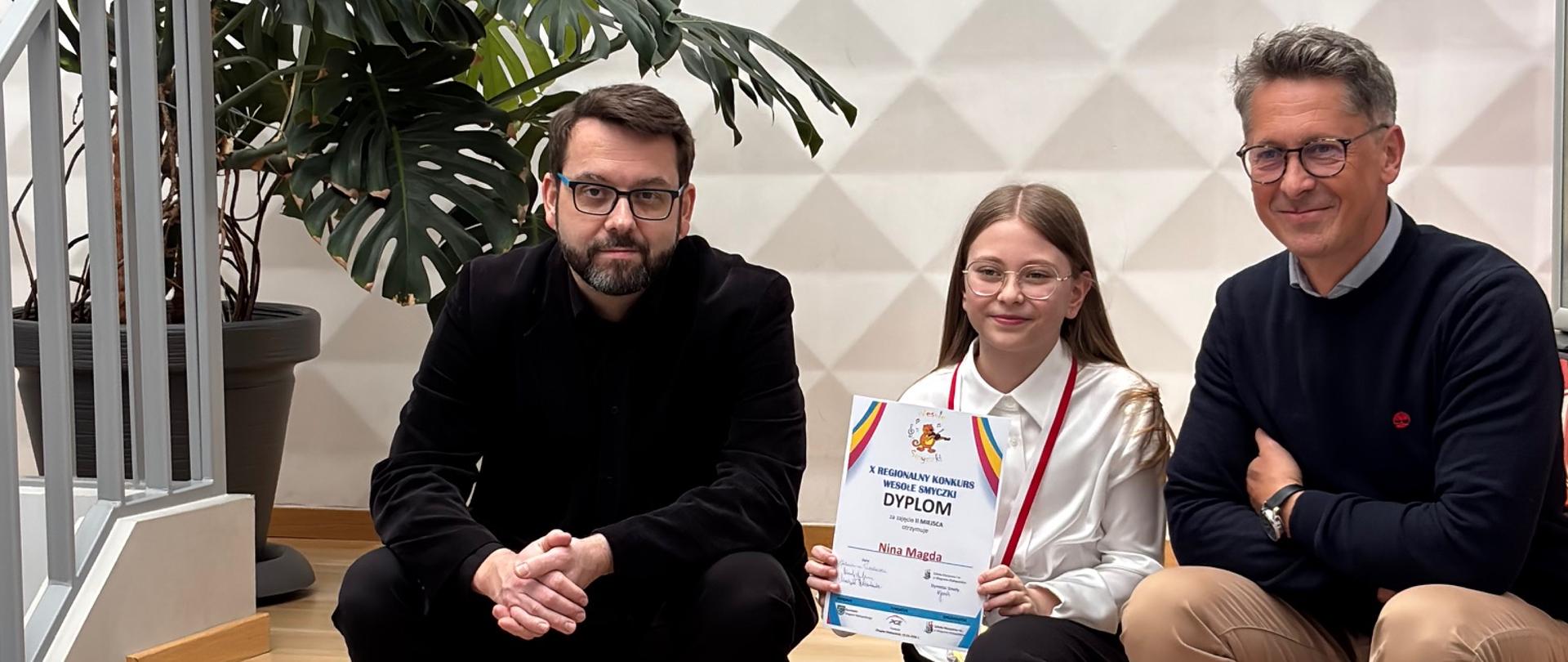 Uczennica siedząca z dyplomem obok dwóch nauczycieli podczas X Regionalnego Konkursu „Wesołe smyczki” w Głogowie Małopolskim.