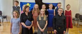 Na zdjęciu grupa uczniów odświętnie ubranych. Uczniowie ustawieni są w dwóch rzędach. Ci z tyłu stoją na scenie. Wśród uczniów stoi ubrana na czarno kobieta brunetka, która trzyma granatową teczkę. Za nimi czarny fortepian.